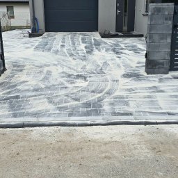 AD-Bruk - Nowoczesny podjazd z szarej kostki brukowej, częściowo zasypanej piaskiem, z bramą wjazdową i ogrodzeniem z szarych bloczków. Widoczne ślady po zagęszczarce.