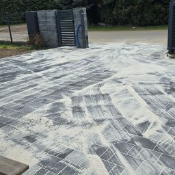 AD-Bruk - Świeżo ułożona, szara kostka brukowa z piaskiem, prowadząca do nowoczesnej, przesuwnej bramy wjazdowej. W tle widoczny traktor i zieleń.