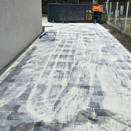 AD-Bruk - Świeżo ułożona, szara kostka brukowa z białym piaskiem na fugach, prowadząca wzdłuż szarej ściany budynku i ogrodzenia z siatki. W tle brama wjazdowa i osoba.