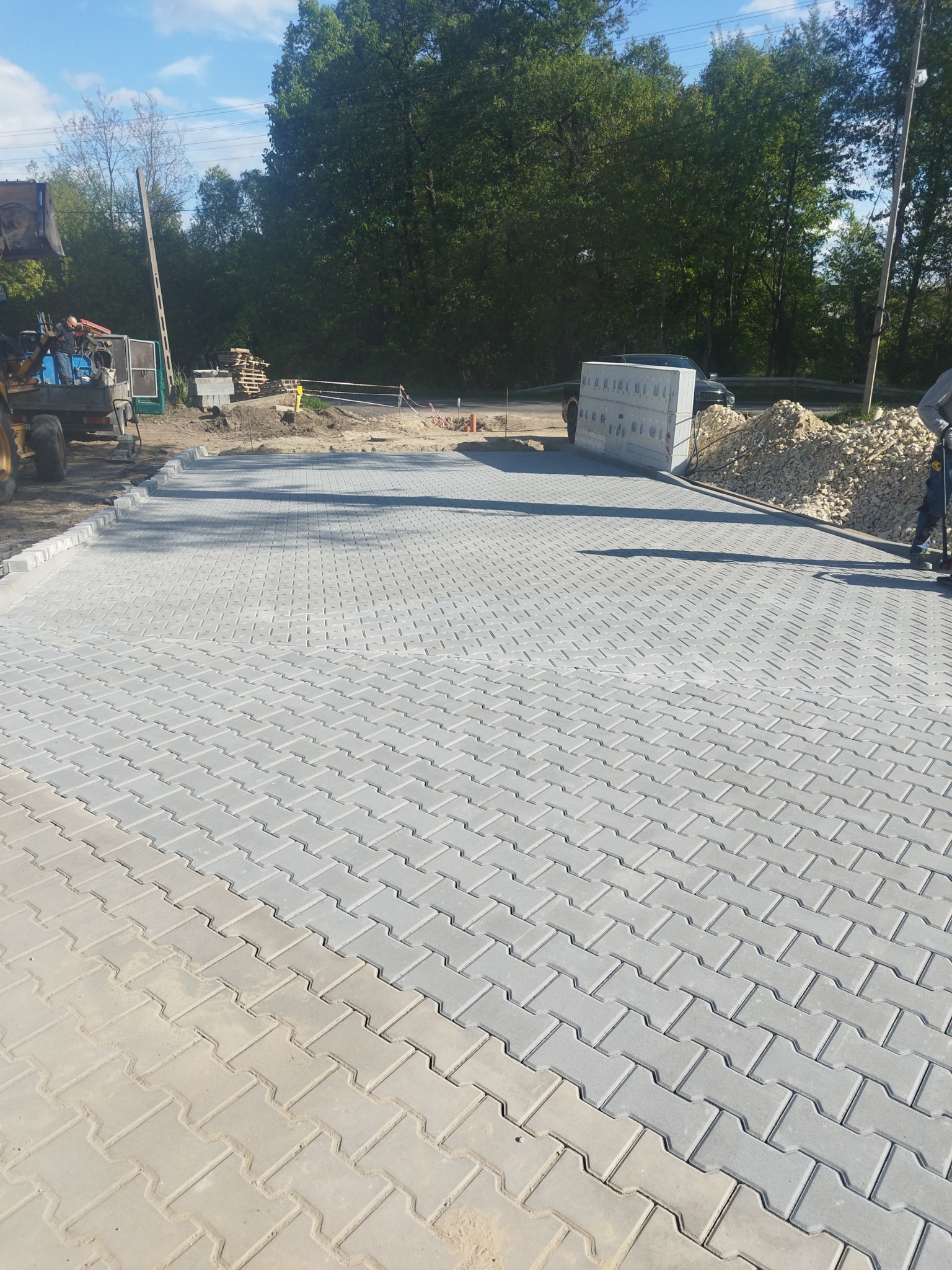 Szara kostka brukowa ułożona w jodełkę, widoczny fragment placu budowy z koparką i stertą kamieni. Częściowo ukończona nawierzchnia.