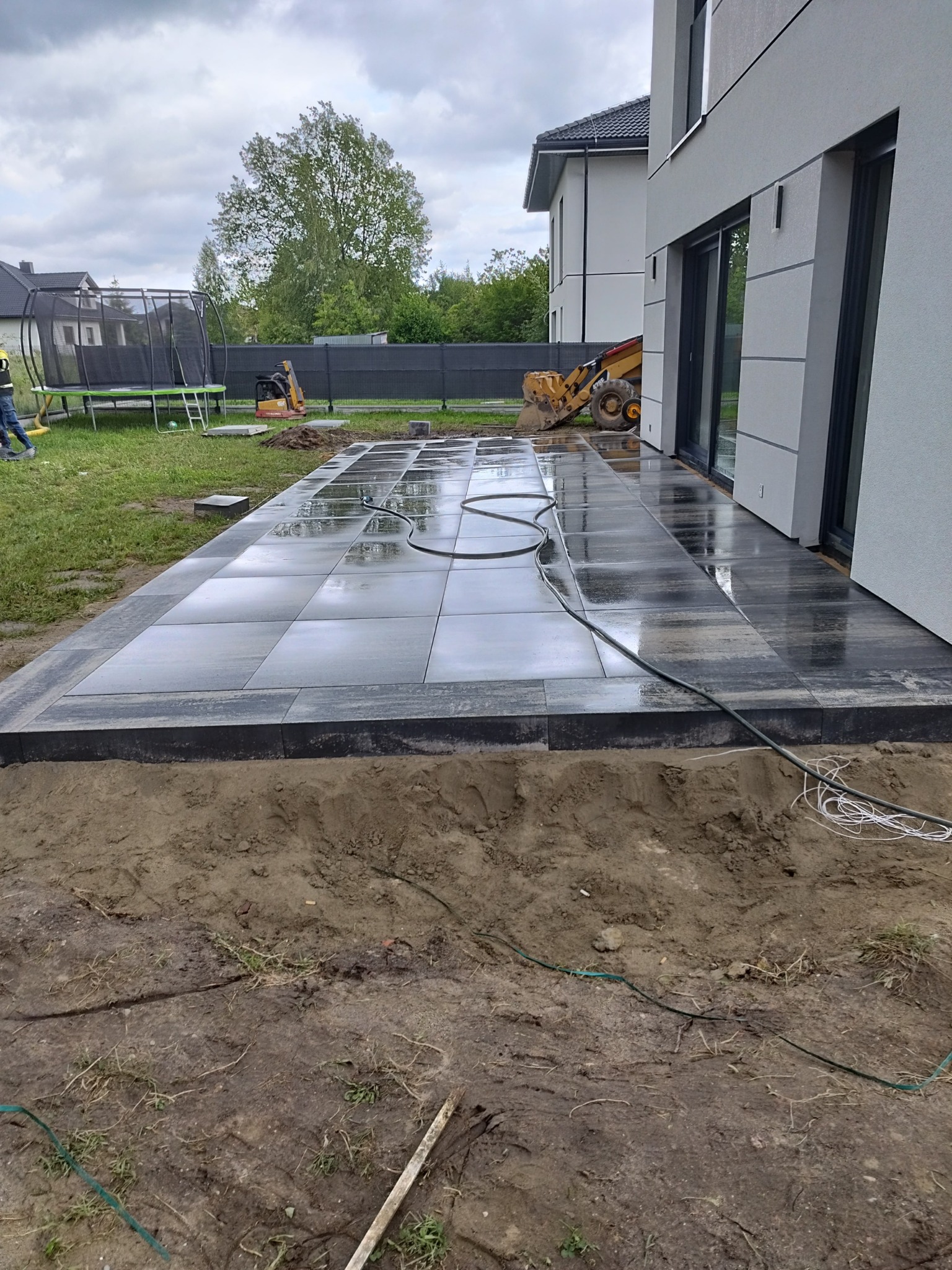 Nowoczesny taras z szarych płyt, częściowo ułożony, z widocznym wykopem i sprzętem budowlanym w tle. Świeżo ułożona nawierzchnia po deszczu.