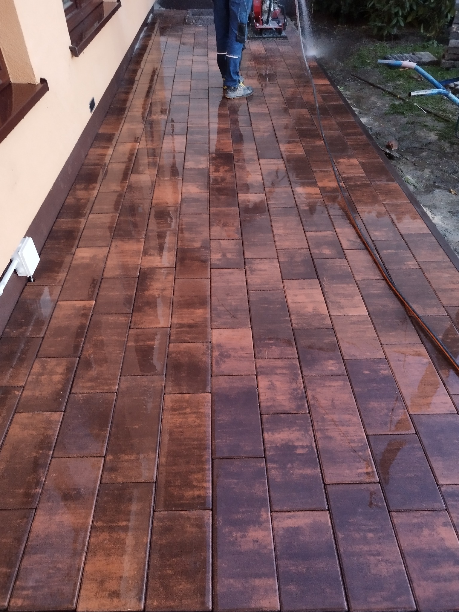 Ciemnobrązowa kostka brukowa ułożona przy budynku, czyszczona myjką ciśnieniową. Widoczne mokre kostki i fragment osoby obsługującej urządzenie.