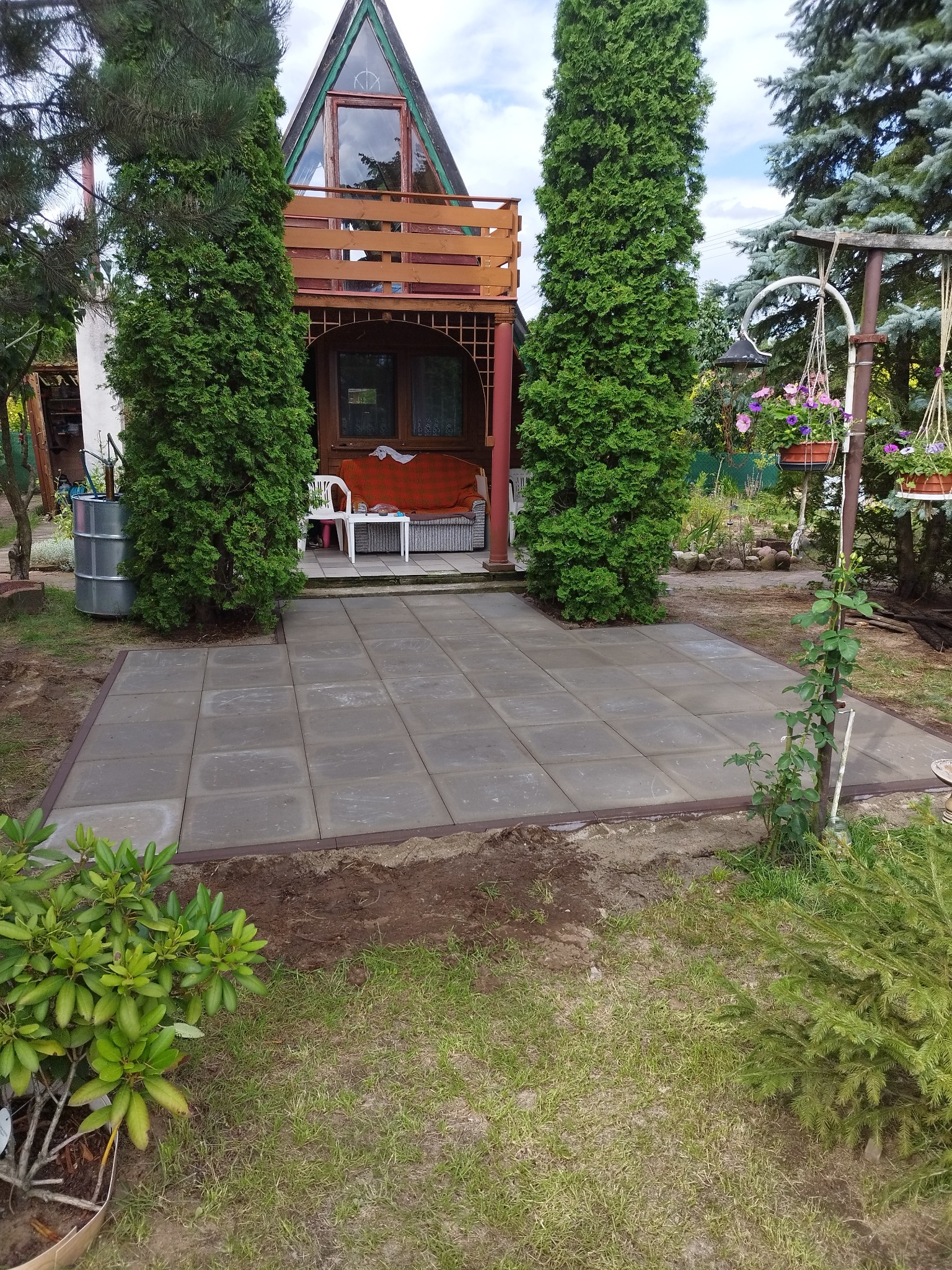 Ułożona, szara kostka brukowa tworząca patio przed domem z balkonem, otoczona zielenią w Łodzi. Widoczne elementy ogrodu i fragment budynku.