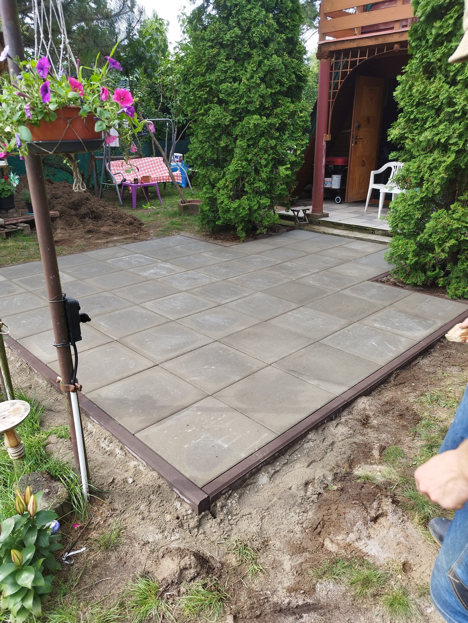 Kwadratowa, szara kostka brukowa z brązowym obrzeżem, ułożona na ziemi w ogrodzie. W tle roślinność i elementy architektury ogrodowej.