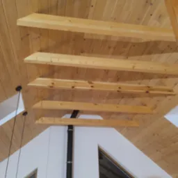 Drewniany sufit z belkami w jasnym pomieszczeniu z oknami. Widoczne elementy oświetlenia i komina. Wykończenie wnętrza w stylu skandynawskim z naturalnych materiałów.