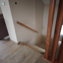 GM Vector plus - Wnętrze domu z drewnianymi schodami i balustradą. Widoczne drewniane stopnie, poręcz oraz słupki balustrady w ciepłym odcieniu brązu. Podłoga wyłożona panelami imitującymi drewno.