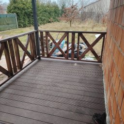 GM Vector plus - Zadaszony taras z brązową deską kompozytową i drewnianą balustradą w kształcie krzyży. Widok na ogród zza balustrady. Częściowo widoczna ściana budynku z cegły.