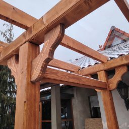 GM Vector plus - Zdobiona, impregnowana konstrukcja z drewna, prawdopodobnie pergola, z widocznymi detalami wykończenia i elementami dachu w tle, uchwycona w pochmurny dzień.