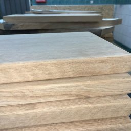 GM Vector plus - Stos jasnych, drewnianych desek o gładkiej powierzchni, ułożonych jedna na drugiej. Widoczne słoje drewna i krawędzie desek. W tle inne elementy drewniane i fragment ściany.