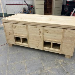 GM Vector plus - Surowa, jasna konstrukcja z drewna, przypominająca skrzynię z otworami, ustawiona na szarej podłodze z płytek. Widoczne niedoskonałości i surowy stan wykończenia.