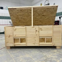 GM Vector plus - Surowa, drewniana konstrukcja skrzyni z otwieranym wiekiem z płyty OSB, widoczna w warsztacie. Skrzynia na czterech nogach, z otworami w dolnej części. W tle narzędzia.