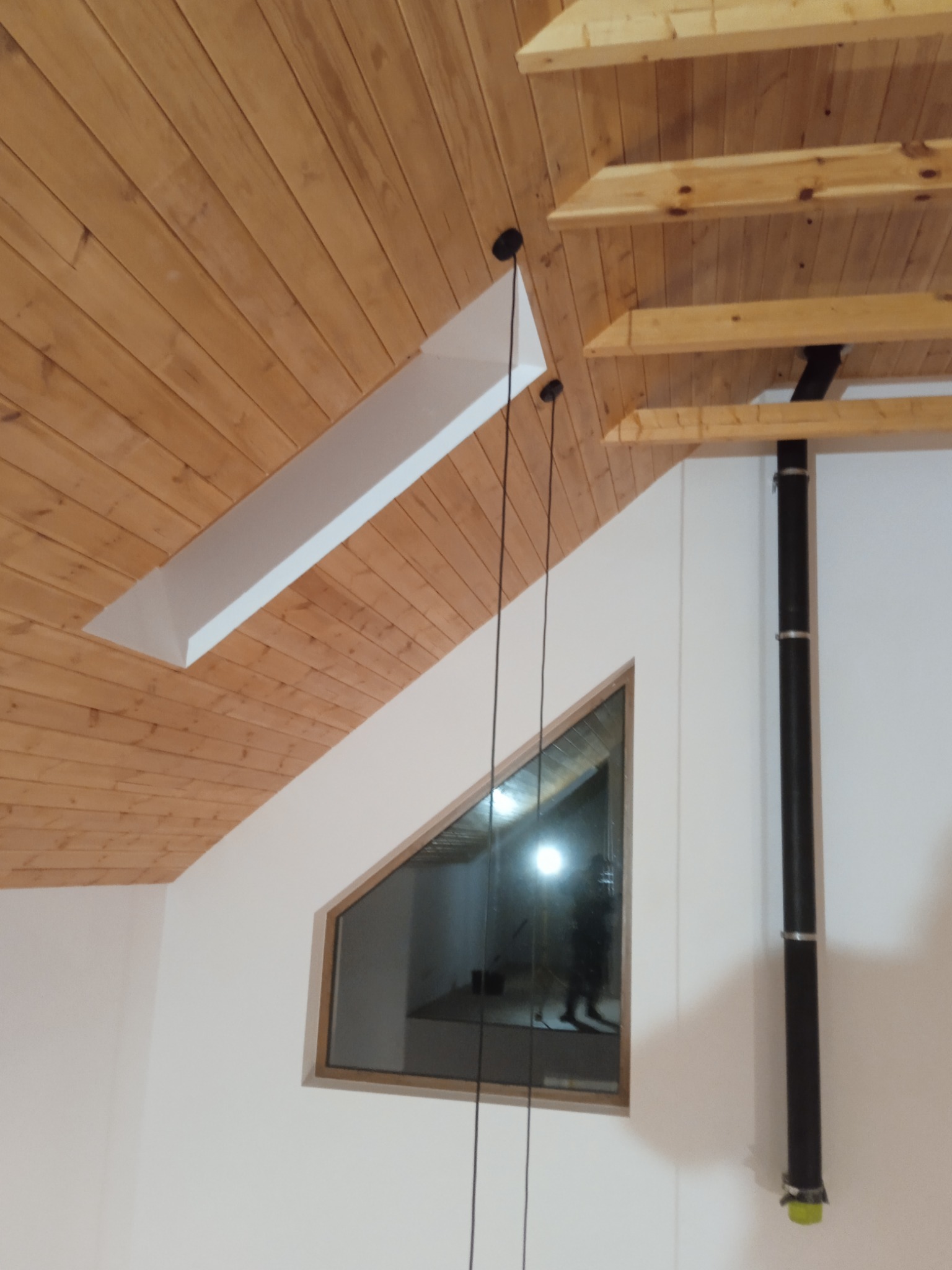 Drewniana boazeria na poddaszu z oknem w kształcie trójkąta i czarną rurą. Widoczne elementy instalacji oświetleniowej i wykończenia sufitu.
