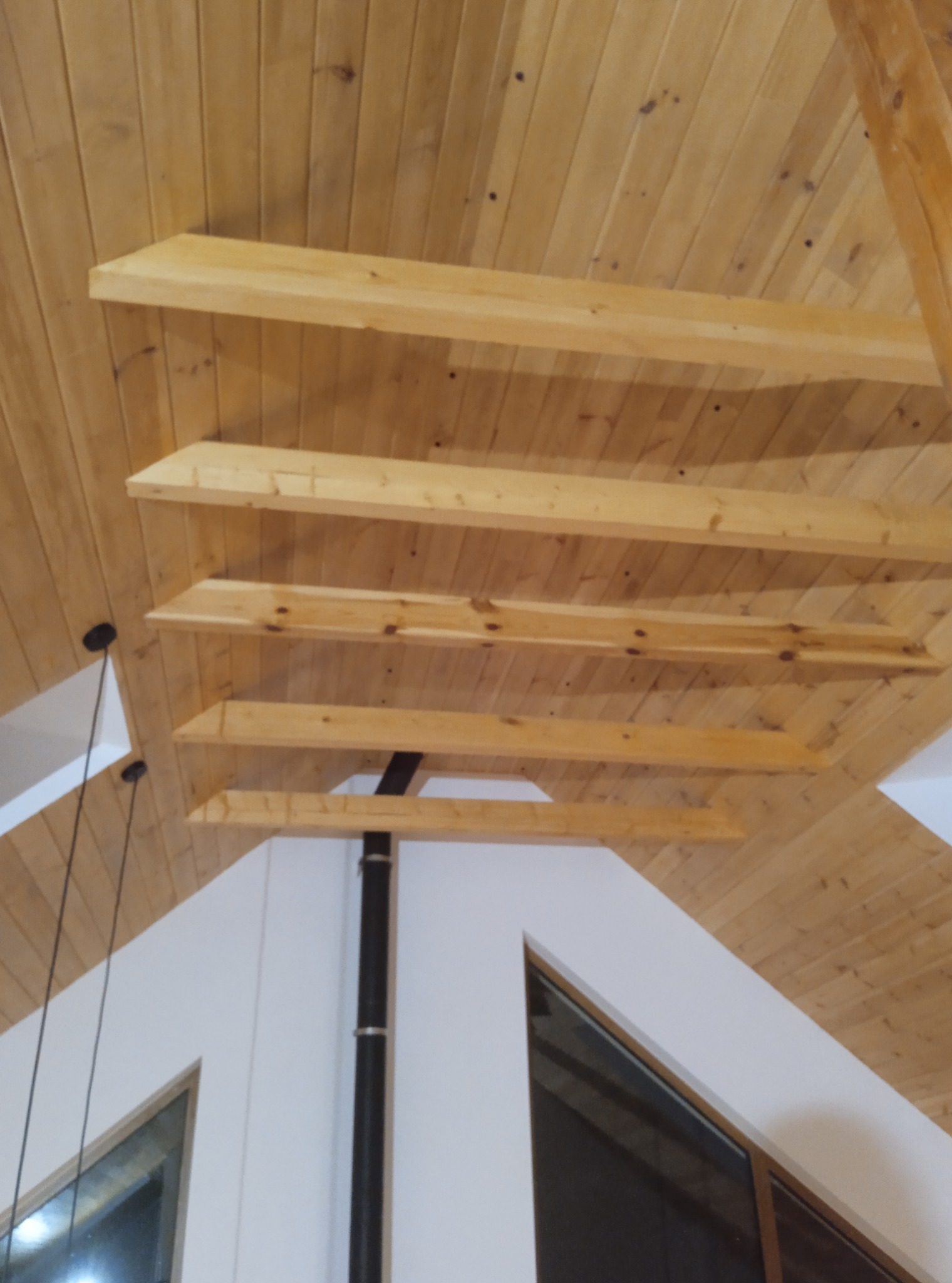 Drewniany sufit z belkami w jasnym pomieszczeniu z oknami. Widoczne elementy oświetlenia i komina. Wykończenie wnętrza w stylu skandynawskim z naturalnych materiałów.