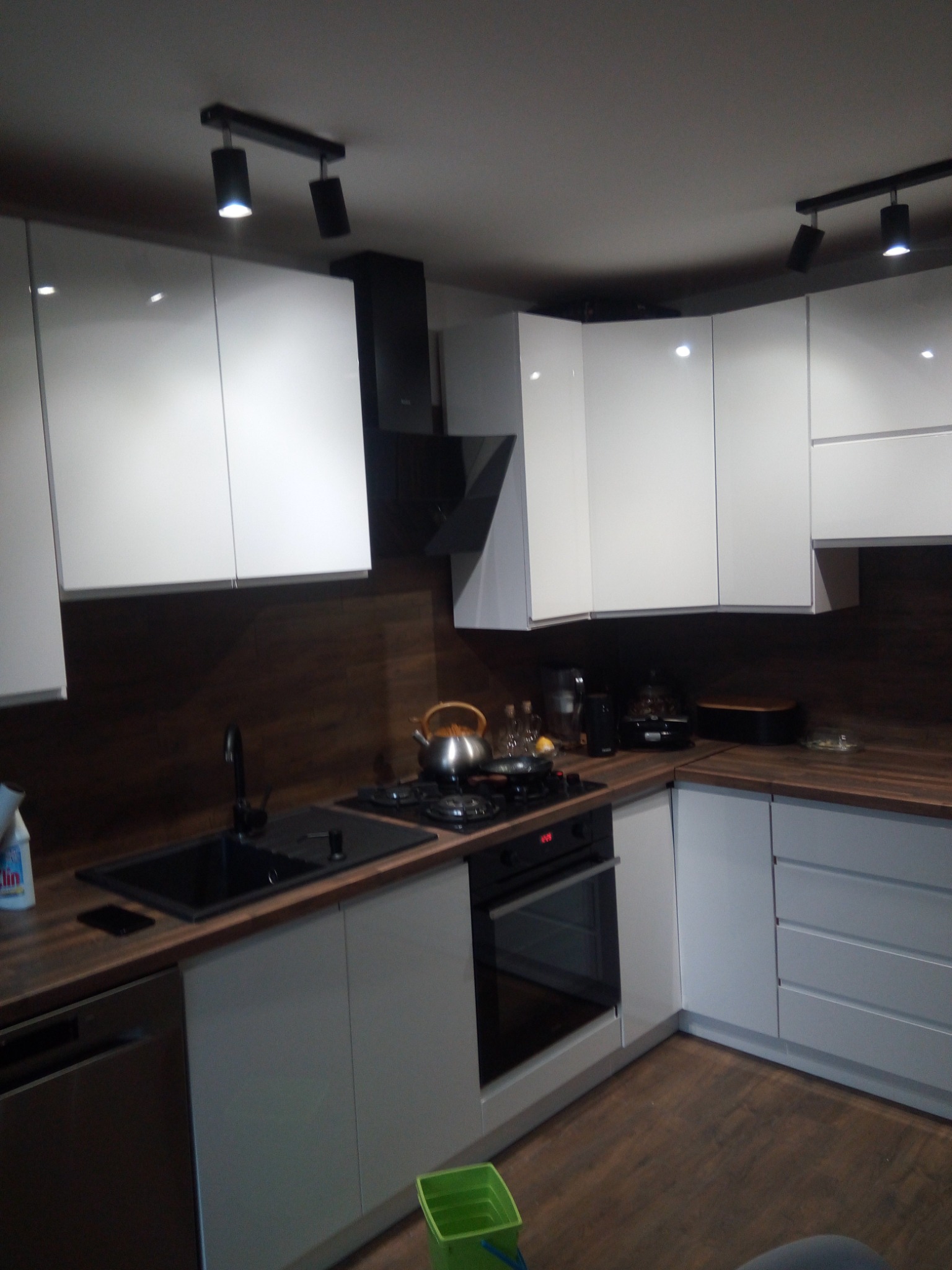 Nowoczesna kuchnia z białymi szafkami, drewnianym blatem i czarnymi akcentami, w tym zlewozmywakiem i oświetleniem punktowym. Widoczny fragment podłogi z paneli.