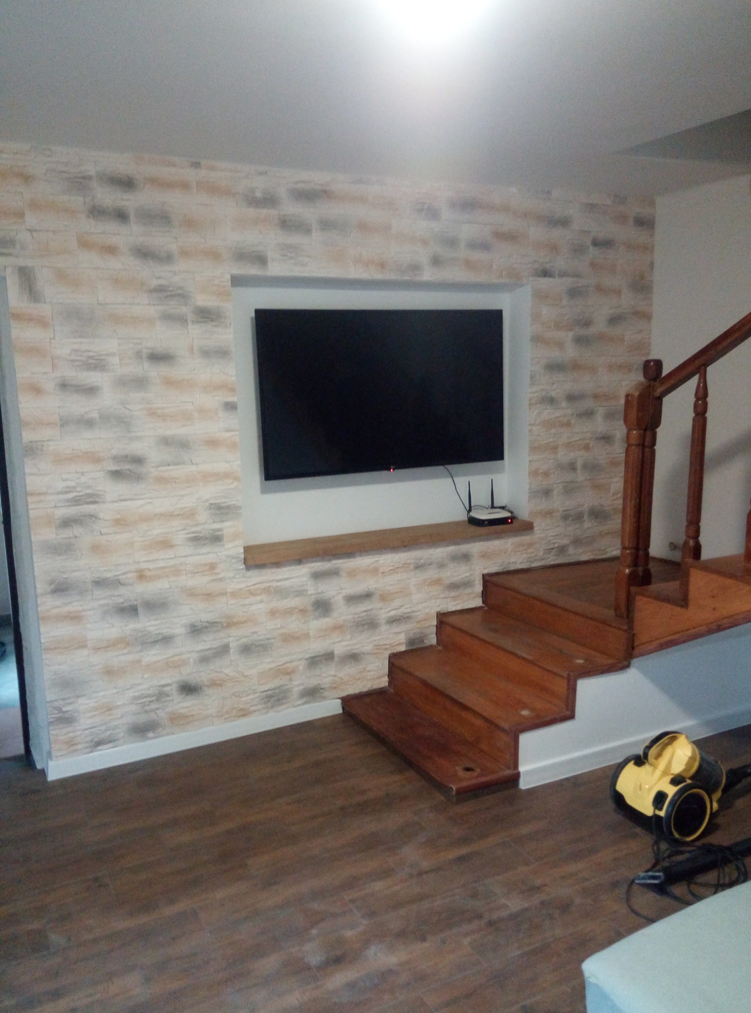 Ściana z dekoracyjną okładziną imitującą cegłę, wnęka na telewizor z półką, drewniane schody i parkiet. Widoczny odkurzacz.
