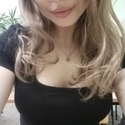 Portret młodej kobiety z blond włosami, noszącej czarną bluzkę i złoty naszyjnik z sercem, wykonany w pomieszczeniu z biurkiem i rośliną w tle.