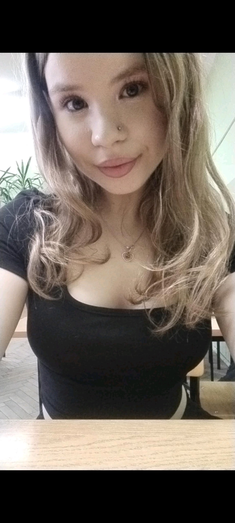 Portret młodej kobiety z blond włosami, noszącej czarną bluzkę i złoty naszyjnik z sercem, wykonany w pomieszczeniu z biurkiem i rośliną w tle.