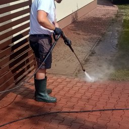 Pure House Marta Wielgomasz - Mężczyzna w gumowych butach czyści czerwoną kostkę brukową myjką ciśnieniową. W tle budynek i bus.