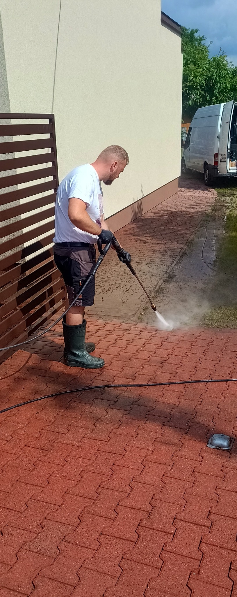 Mężczyzna w gumowych butach czyści czerwoną kostkę brukową myjką ciśnieniową. W tle budynek i bus.