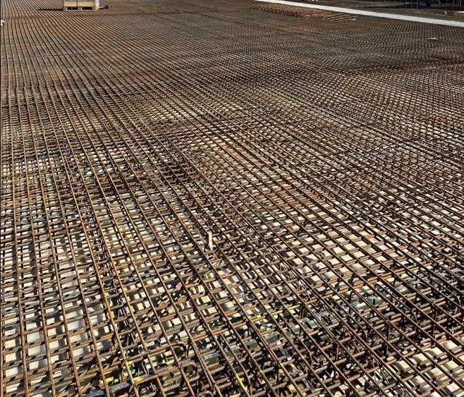 Siatka zbrojeniowa ułożona na podłożu, przygotowana do zalania betonem. Widok z góry, ukazujący regularny wzór stalowych prętów. Elementy szalunku w tle.