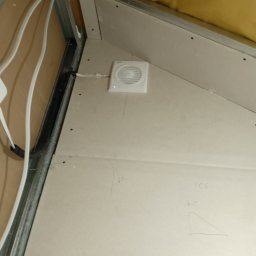 Konrad Mikuli - Skośne poddasze z płytami karton-gips, widoczny wentylator, metalowe profile, instalacja elektryczna i izolacja. Na płytach widoczne odręczne notatki.