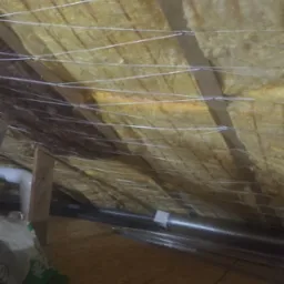 Ocieplenie poddasza wełną mineralną, widoczne drewniane belki i białe sznurki mocujące izolację. Fragment instalacji wentylacyjnej i podłoga z płyt OSB.