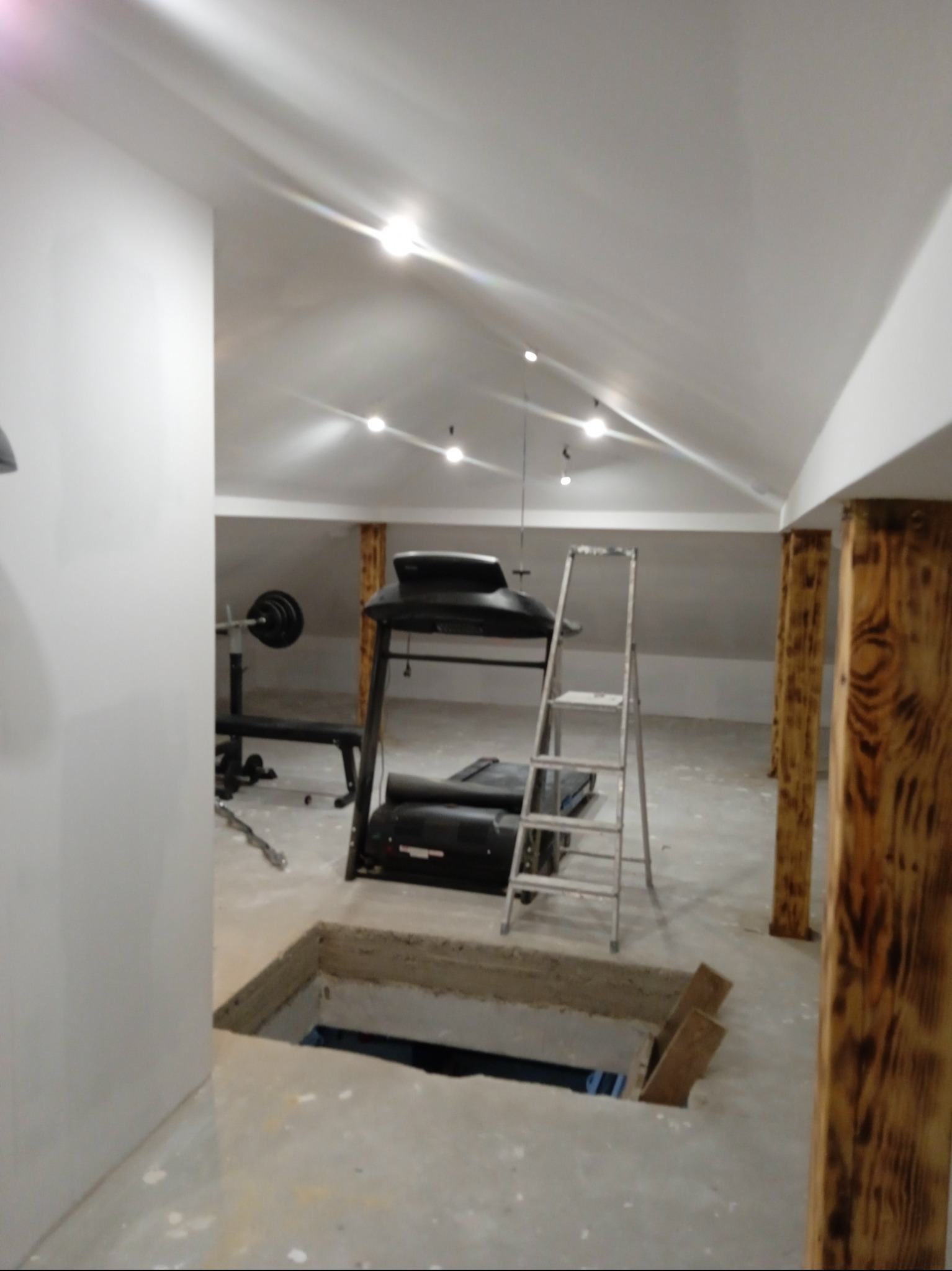 Adaptacja poddasza na siłownię: widok na sufit z oświetleniem, sprzęt do ćwiczeń, drewniane słupy i otwór w podłodze z drabinką.