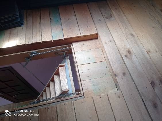 Drewniane wykończenie poddasza z otworem na schody strychowe, widoczne mechanizmy sprężynowe. Ujęcie z dołu, surowe drewno, widoczne słoje i detale konstrukcji.