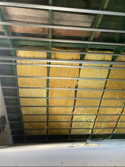 Ocieplenie poddasza wełną mineralną, widok od dołu. Żółta wełna między drewnianymi belkami i metalowymi profilami. Częściowo widoczne kable.