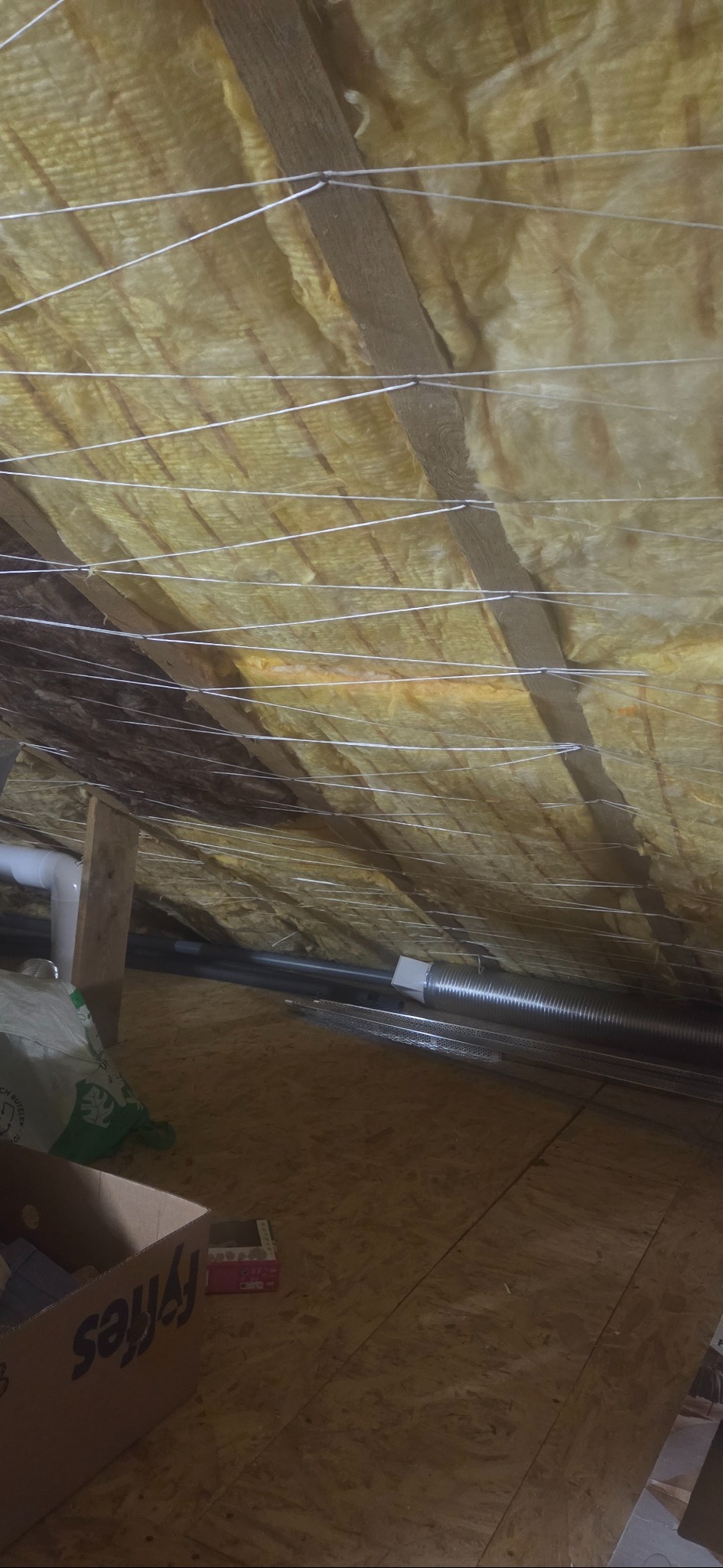 Ocieplenie poddasza wełną mineralną, widoczne drewniane belki i białe sznurki mocujące izolację. Fragment instalacji wentylacyjnej i podłoga z płyt OSB.
