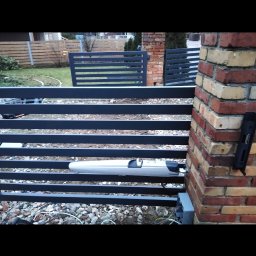 Serhii Kuzmych - Szara brama lamelowa z automatyką, widok z bliska na mechanizm i słupek z cegły. Na tle trawnik i ogrodzenie posesji.