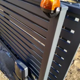 Nowoczesna brama przesuwna z automatyką SEA, lampa ostrzegawcza. Szare, poziome panele tworzą minimalistyczny design. Widoczne elementy montażowe i napęd bramy.