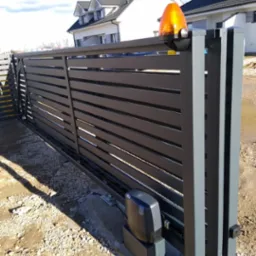 Nowoczesna, przesuwna brama w kolorze grafitowym z automatyką, z widoczną lampą ostrzegawczą. W tle dom z jasną elewacją i szarą dachówką.