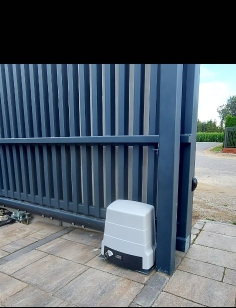 Szara, przesuwna brama z pionowymi panelami i automatyką przy kostce brukowej. Widoczny napęd bramy w jasnej obudowie i fragment zieleni w tle.