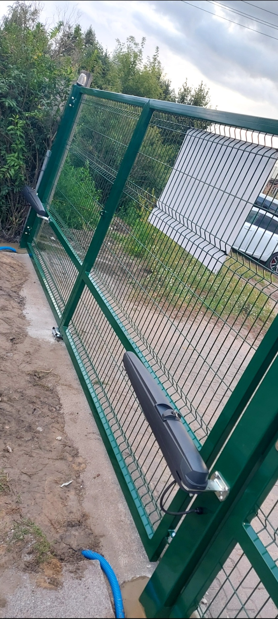 Zielona brama panelowa z automatyką, widok z boku. Widoczne siłowniki. W tle zieleń i fragment białego samochodu. Błękitny wężyk na ziemi.