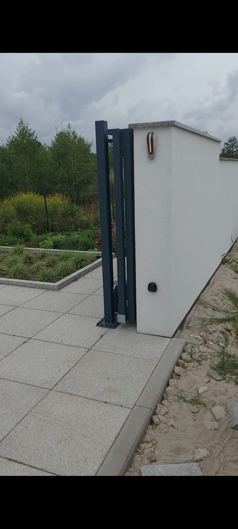 Nowoczesna brama przesuwna w kolorze grafitowym, zintegrowana z białym murem i oświetleniem. Widok na ogród i kostkę brukową.