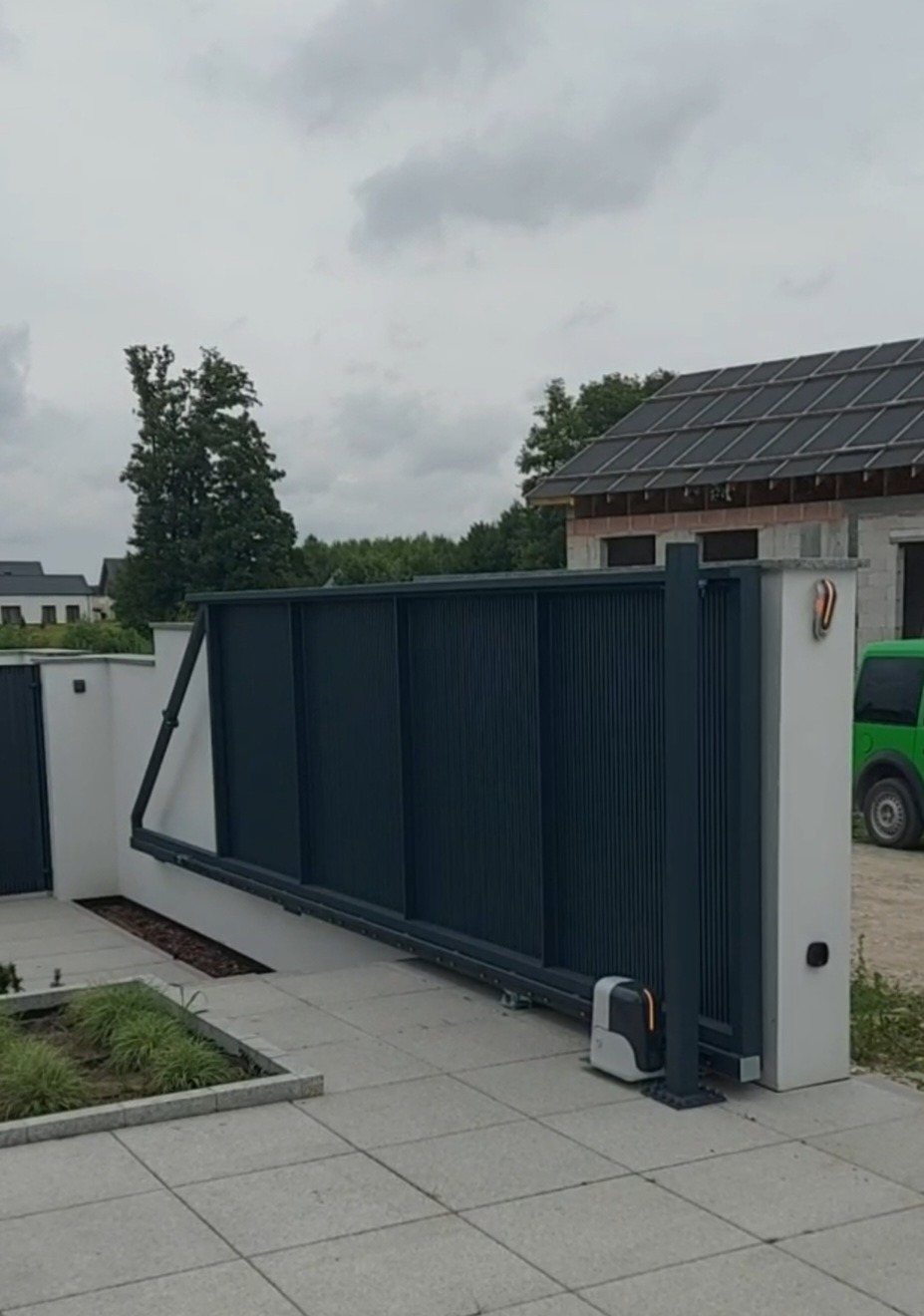 Nowoczesna, przesuwna brama w kolorze grafitowym, z widocznym napędem automatycznym. Brama częściowo otwarta, odsłaniająca fragment posesji i budynek w tle.