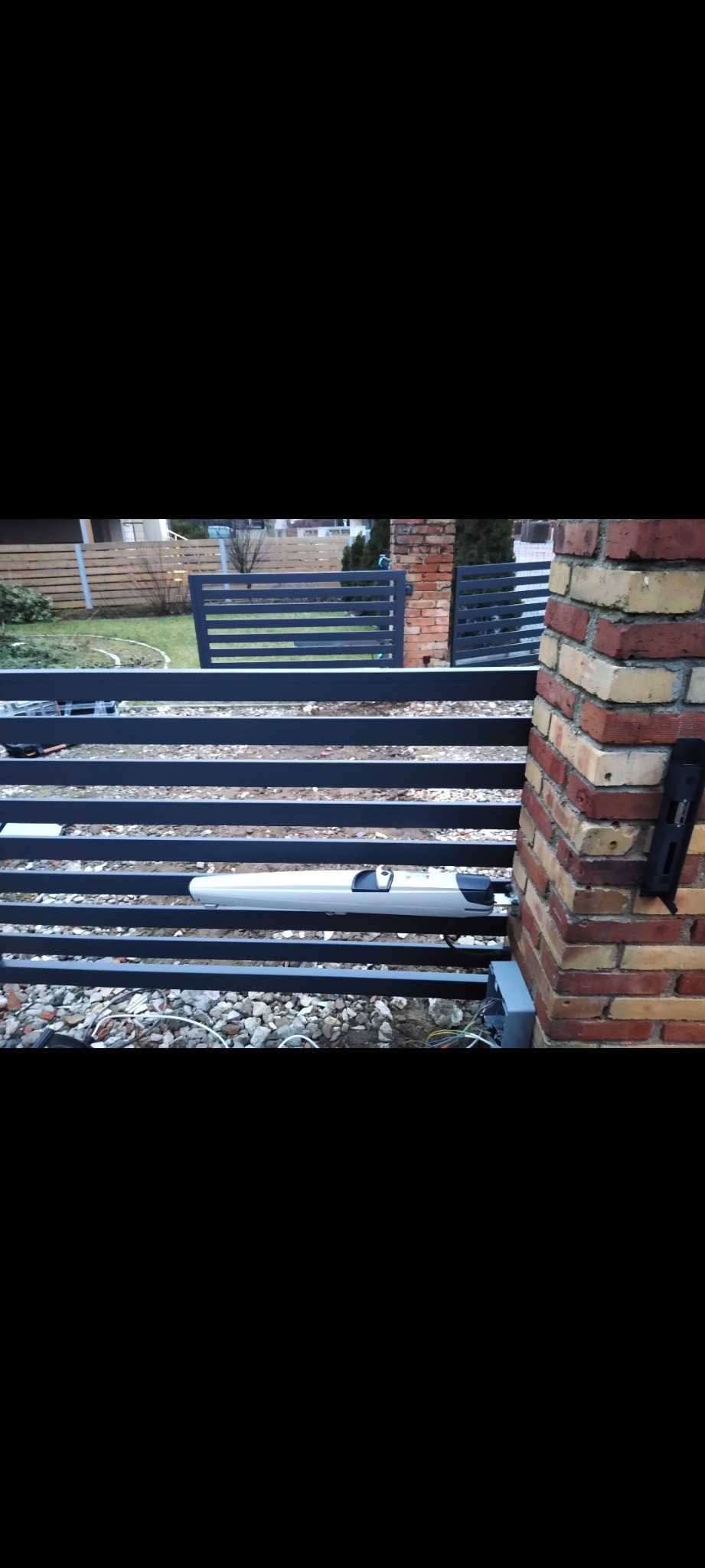 Szara brama lamelowa z automatyką, widok z bliska na mechanizm i słupek z cegły. Na tle trawnik i ogrodzenie posesji.