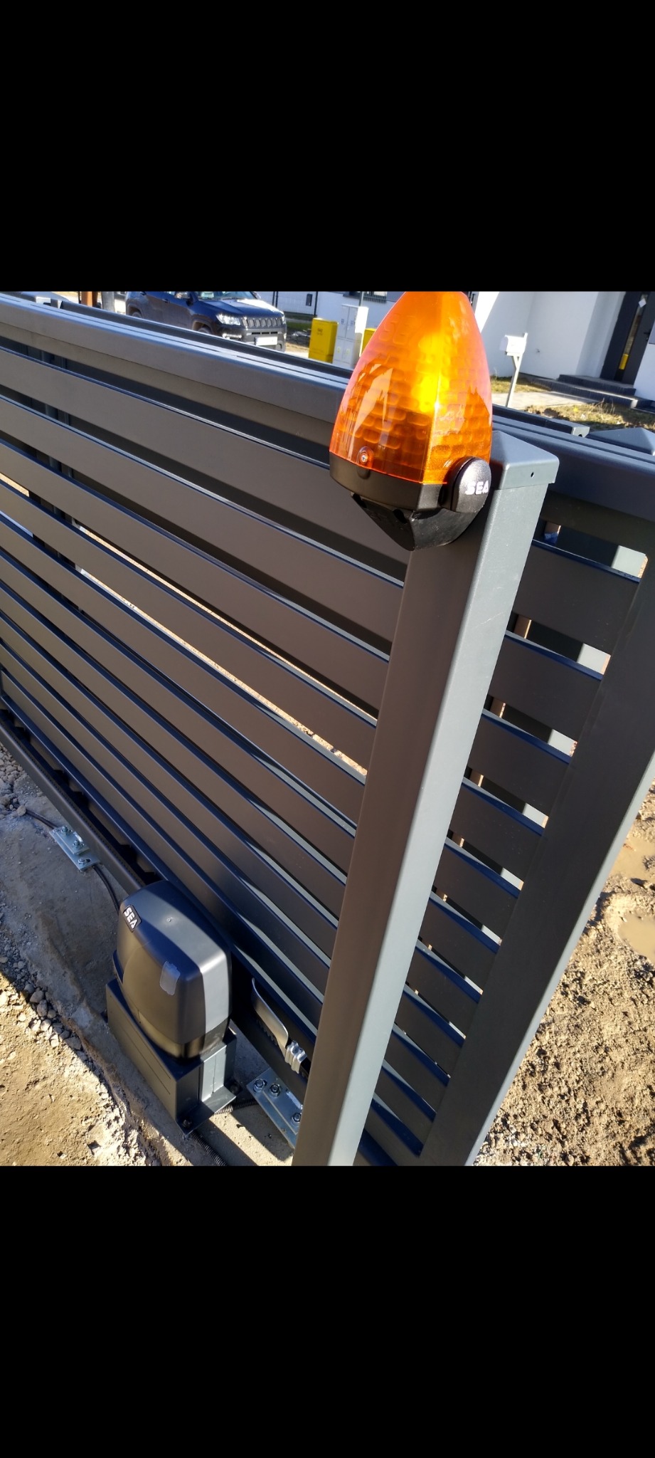 Nowoczesna brama przesuwna z automatyką SEA, lampa ostrzegawcza. Szare, poziome panele tworzą minimalistyczny design. Widoczne elementy montażowe i napęd bramy.