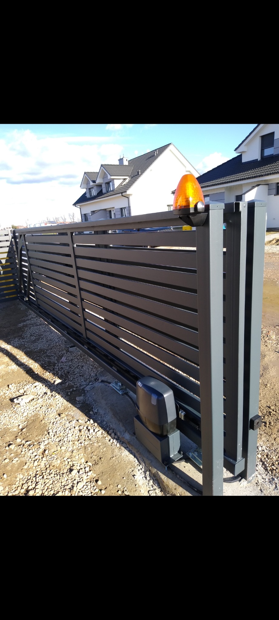 Nowoczesna, przesuwna brama w kolorze grafitowym z automatyką, z widoczną lampą ostrzegawczą. W tle dom z jasną elewacją i szarą dachówką.