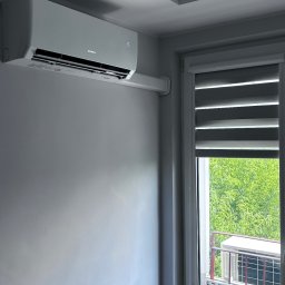 Ecotherm - Biały klimatyzator Gree na ścianie w jasnym pokoju, widok przez okno na jednostkę zewnętrzną na balkonie. Minimalistyczny design wnętrza.