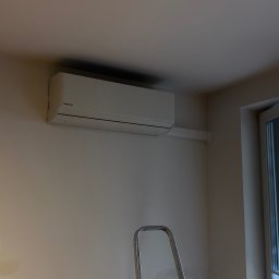 Ecotherm - Biały klimatyzator Panasonic na ścianie w jasnym pokoju, widoczna drabina i otwarte drzwi balkonowe. Nowoczesny design, świeżo po montażu, gniazdka elektryczne na ścianie.