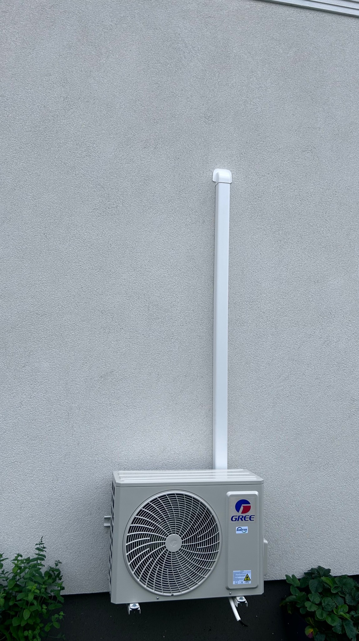 Zamontowany klimatyzator marki Gree na szarej, teksturowanej ścianie budynku. Widoczna biała rura odprowadzająca. U dołu roślinność.