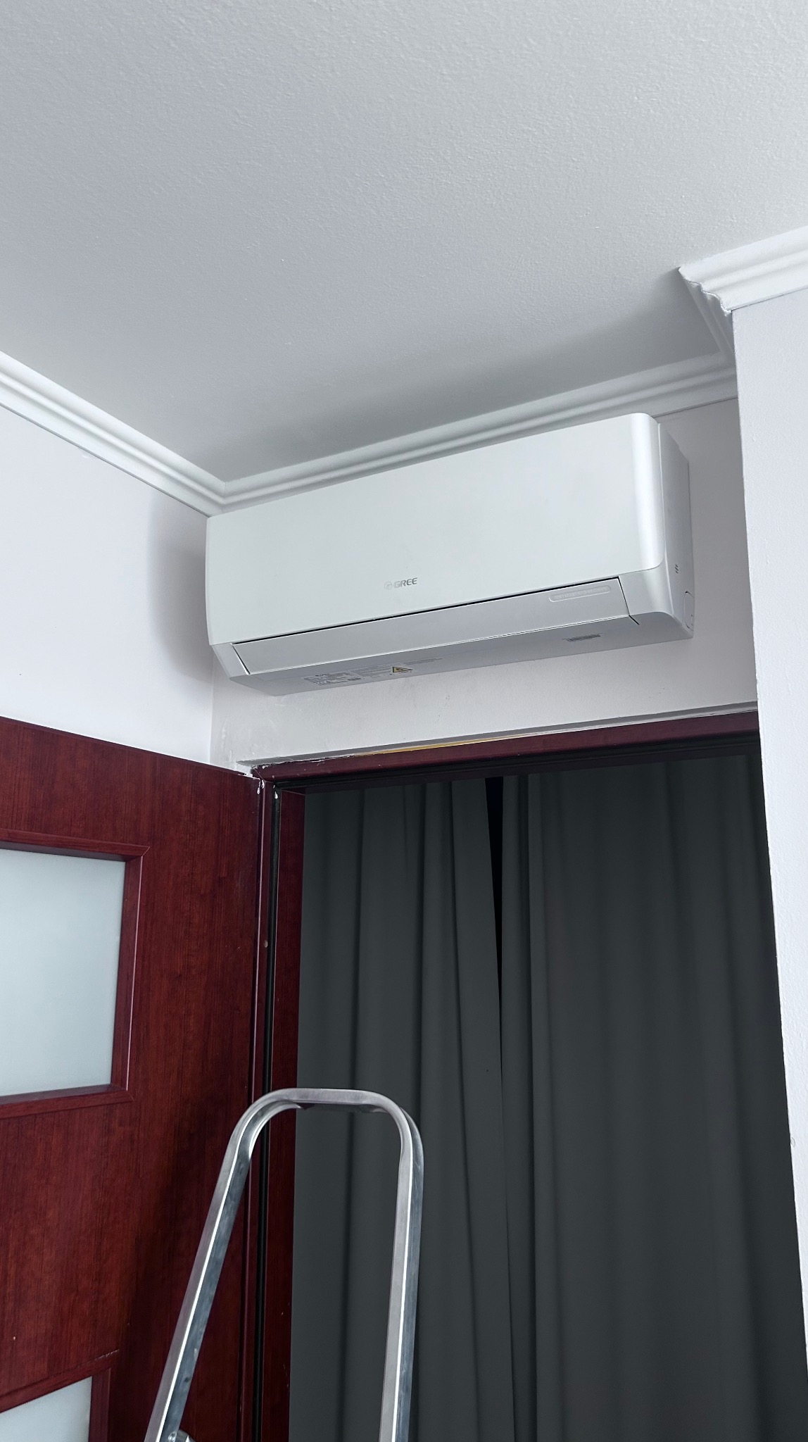 Nowoczesny, biały klimatyzator marki Gree zamontowany nad drzwiami z zasłoniętym oknem, widziany z dołu. Fragment drabiny sugeruje montaż lub serwis.