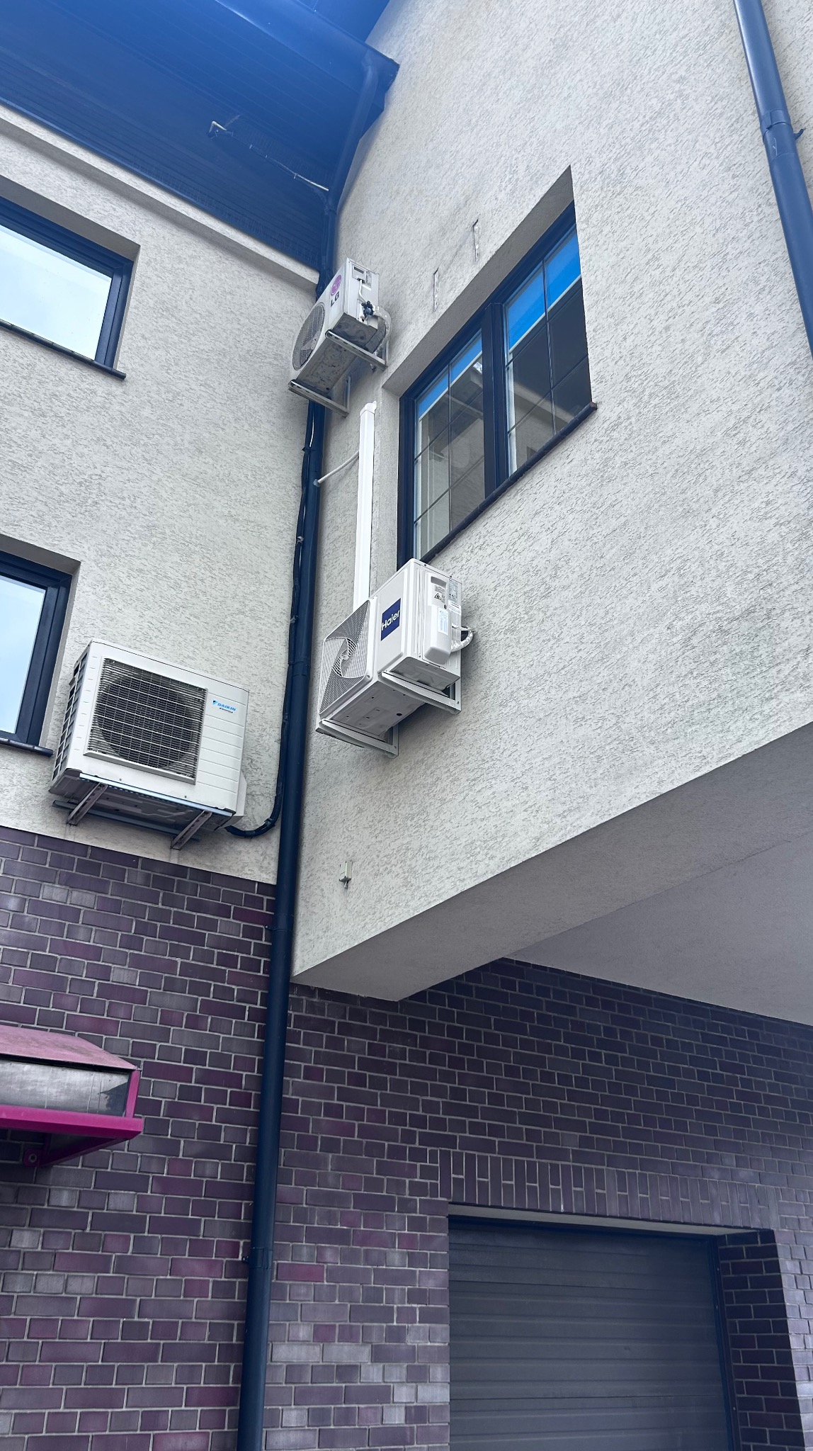 Montaż klimatyzacji: trzy jednostki zewnętrzne różnych marek (Daikin, Haier, LG) na elewacji budynku z cegły i tynku strukturalnego. Rury odprowadzające skropliny.