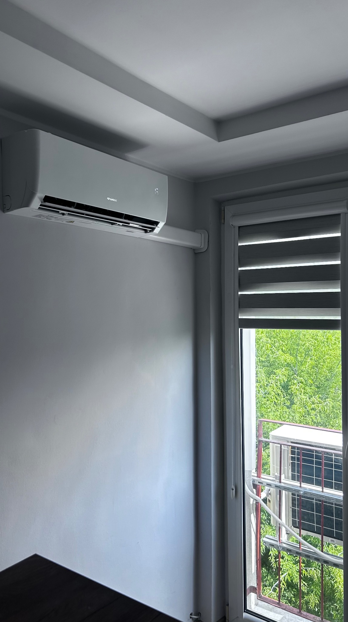 Biały klimatyzator Gree na ścianie w jasnym pokoju, widok przez okno na jednostkę zewnętrzną na balkonie. Minimalistyczny design wnętrza.