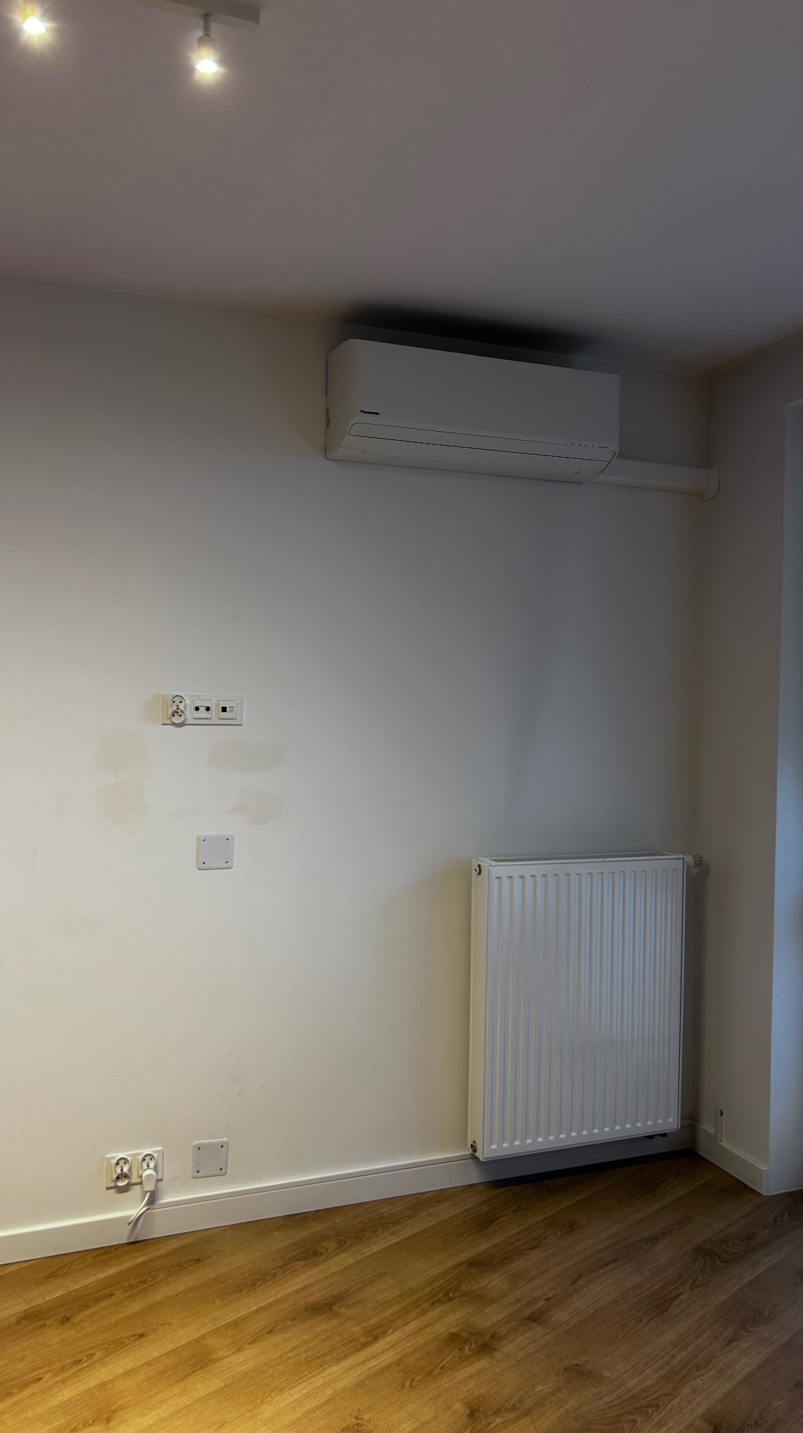 Biały klimatyzator Panasonic na ścianie nad grzejnikiem w jasnym pokoju z drewnianą podłogą. Widoczne gniazdka elektryczne i oświetlenie sufitowe.