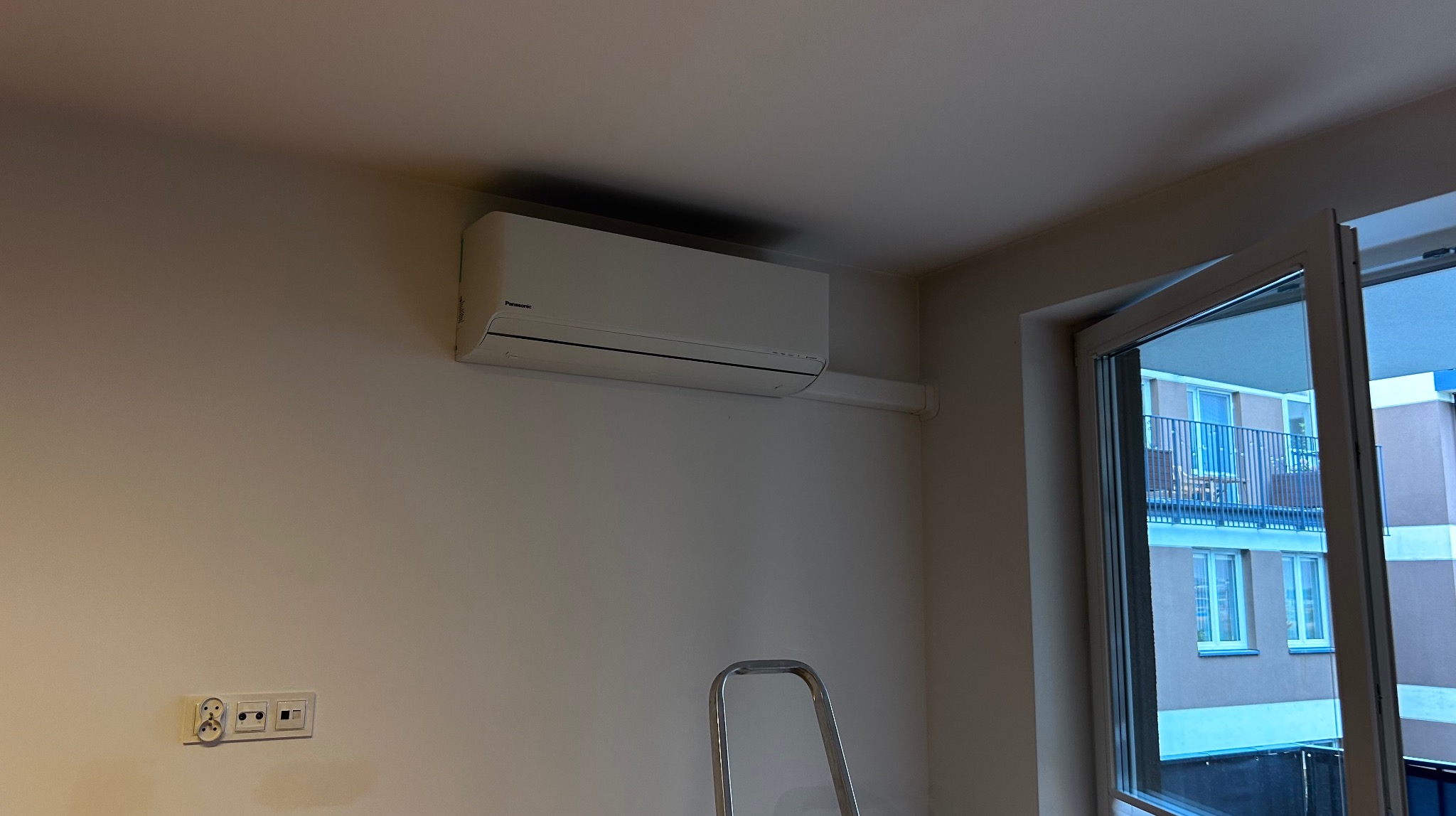 Biały klimatyzator Panasonic na ścianie w jasnym pokoju, widoczna drabina i otwarte drzwi balkonowe. Nowoczesny design, świeżo po montażu, gniazdka elektryczne na ścianie.