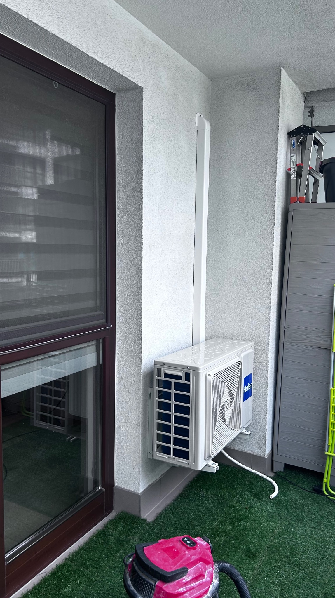 Biały klimatyzator Haier zamontowany na ścianie balkonu z zieloną sztuczną trawą i odkurzaczem w rogu. Widoczna drabina i szafka.
