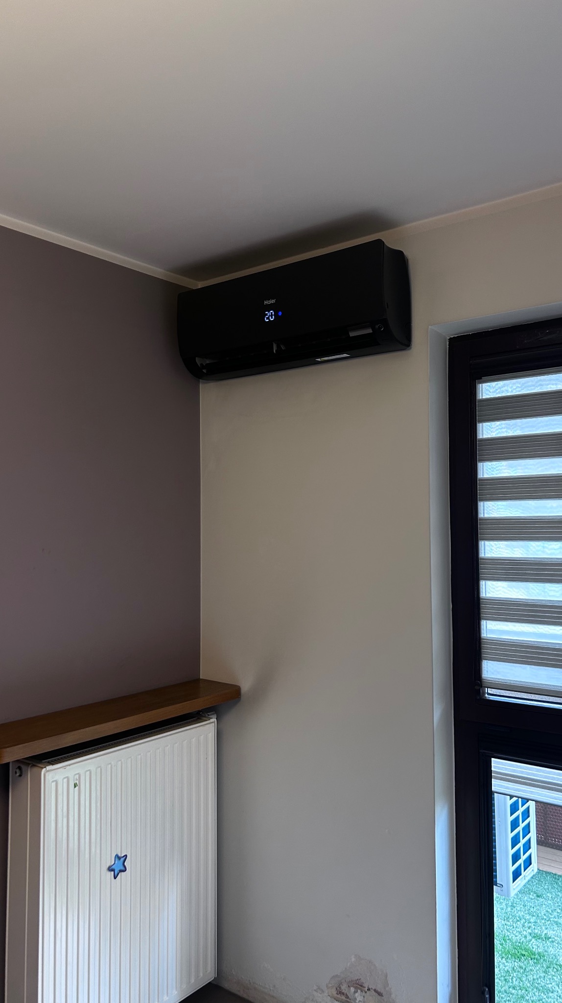Czarny klimatyzator Haier na ścianie w pokoju, obok okna z żaluzjami. Pod klimatyzatorem grzejnik z półką. Widoczny fragment ściany w dwóch odcieniach.